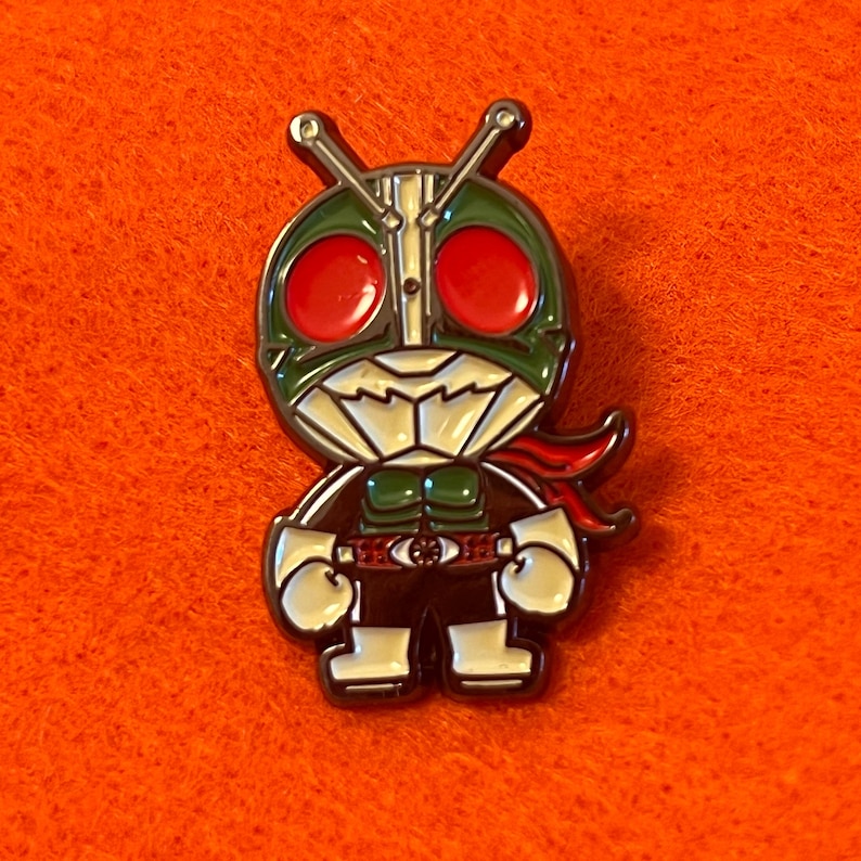 May include: A colorful enamel pin featuring a stylized character with a green helmet, red eyes, and a red scarf. The character is wearing a black and white outfit with a silver belt. The pin is set against an orange background.