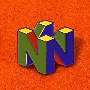 May include: A colorful enamel pin featuring the iconic Nintendo 64 logo. The pin has a 3D design with the letters "N" in green and blue, with red accents and yellow highlights. The pin is set against an orange background.