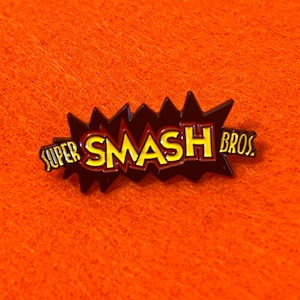 May include: A Super Smash Bros. pin with a black and red burst design. The text "SUPER SMASH BROS." is in yellow with a red outline. The pin is set against an orange background.