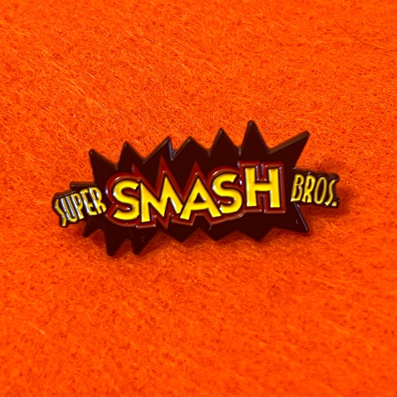 May include: A Super Smash Bros. pin with a black and red burst design. The text "SUPER SMASH BROS." is in yellow with a red outline. The pin is set against an orange background.