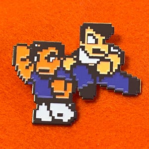 May include: Pixelated enamel pin featuring two figures in a fighting stance. The figures are rendered in a retro 8-bit style, with a color palette of blue, orange, black, and white. The pin is set against an orange background.