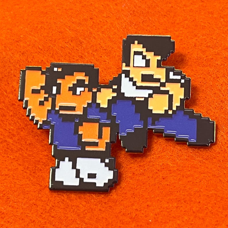 May include: Pixelated enamel pin featuring two figures in a fighting stance. The figures are rendered in a retro 8-bit style, with a color palette of blue, orange, black, and white. The pin is set against an orange background.
