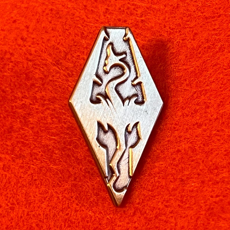 May include: A silver and gold-toned diamond-shaped pin featuring a stylized dragon and two stylized figures. The pin is set against a vibrant red background. The design is intricate and detailed, with a vintage aesthetic.