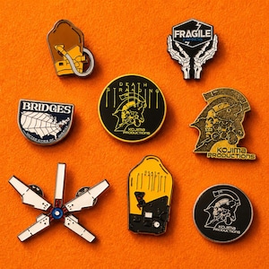 May include: A collection of enamel pins featuring designs from the video game Death Stranding. The pins include logos, character silhouettes, and the text "BRIDGES" and "FRAGILE EXPRESS". The pins are set against an orange background.