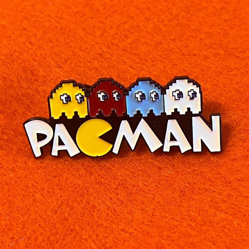 May include: A colorful Pac-Man enamel pin featuring the game's iconic characters: yellow, red, blue, and white ghosts above the word "PACMAN" in white letters. The pin is set against an orange background.