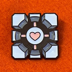 May include: A black, white, gray, and pink enamel pin featuring a cube design with a heart in the center. The pin has a black border and is set against an orange background. The cube design has a 3D effect.