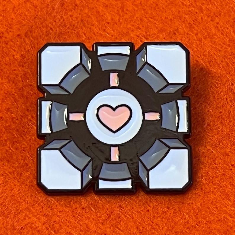 May include: A black, white, gray, and pink enamel pin featuring a cube design with a heart in the center. The pin has a black border and is set against an orange background. The cube design has a 3D effect.