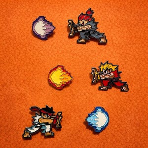 May include: Pixel art enamel pins depicting characters and special moves from a fighting game. The pins show characters in action poses and energy blasts in various colours, set against an orange background.