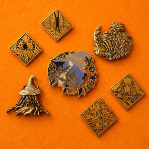 May include: A selection of fantasy-themed enamel pins in gold, black, and blue. The pins depict a witch, a wizard, and other mystical figures. The pins are displayed on an orange surface.