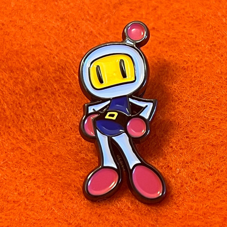 May include: A colorful enamel pin featuring a cartoon character with a yellow face, blue helmet, and pink accents. The character is in a standing pose with arms at its sides. The pin has a black outline and is set against an orange background.