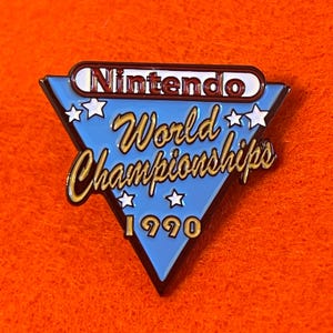 May include: A blue, triangular enamel pin with the text "Nintendo World Championships 1990". The pin features gold lettering and white star accents, with the Nintendo logo at the top. The pin is set against an orange background.