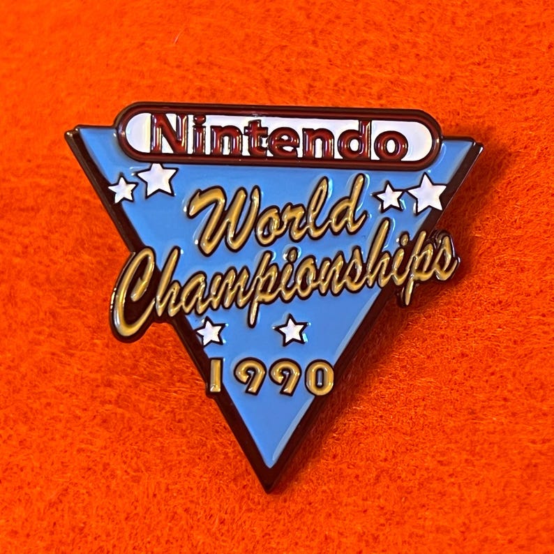 May include: A blue, triangular enamel pin with the text "Nintendo World Championships 1990". The pin features gold lettering and white star accents, with the Nintendo logo at the top. The pin is set against an orange background.