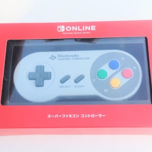 Könnte beinhalten: Ein Nintendo Super Famicom Controller in einer roten Box. Der Controller ist hellgrau mit farbigen Knöpfen und einem Steuerkreuz. Die Box hat das Nintendo Online Logo und japanischen Text.
