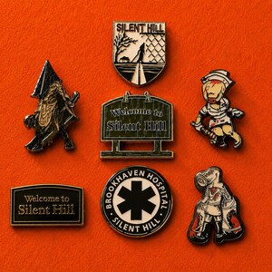 Può includere: Una collezione di sei spille smaltate con immagini di Silent Hill, tra cui una figura di Pyramid Head, un cartello Welcome to Silent Hill, un'infermiera, un emblema del Brookhaven Hospital e una creatura con testa di metallo in bianco e nero.