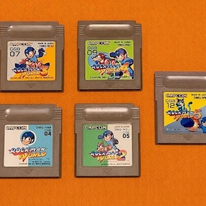 May include: Five grey Capcom Rockman World video game cartridges. Each cartridge has a colourful label with the game title and character art. The cartridges are arranged on an orange surface.