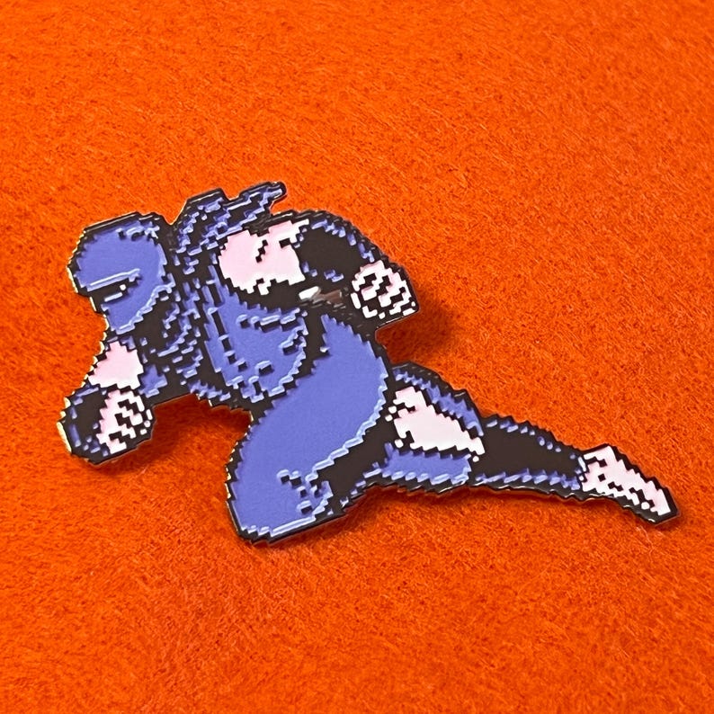May include: A pixelated enamel pin featuring a character in a dynamic pose. The pin has a blue and black color scheme with pink and white accents. The pin is set against an orange background.