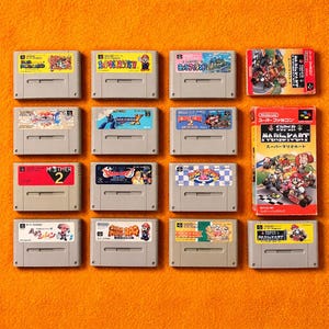 Könnte beinhalten: Eine Auswahl an Super Nintendo (SNES) Spielkassetten und -boxen mit farbenfrohen Etiketten und Kunstwerken. Die Spiele sind auf einer orangefarbenen Oberfläche angeordnet und zeigen Titel wie "Super Mario Kart" und "Mother 2".