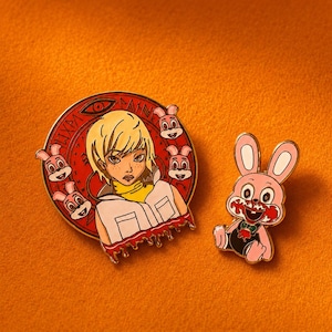 May include: Two enamel pins. The circular pin features a blonde character with a yellow shirt and white vest, surrounded by four pink rabbit heads against a red background with an eye symbol. The second pin is a pink rabbit with a bloody mouth.