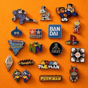 May include: A collection of colorful enamel pins featuring video game and pop culture themes. Designs include characters from Pac-Man, Nintendo, and Atari, along with logos and other iconic imagery. The pins are arranged on an orange surface.