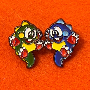 May include: Two enamel pins featuring cartoon dinosaur characters. One is green and yellow, the other is blue and yellow. Both have white eyes, red accents, and are smiling. The pins are set against an orange background.