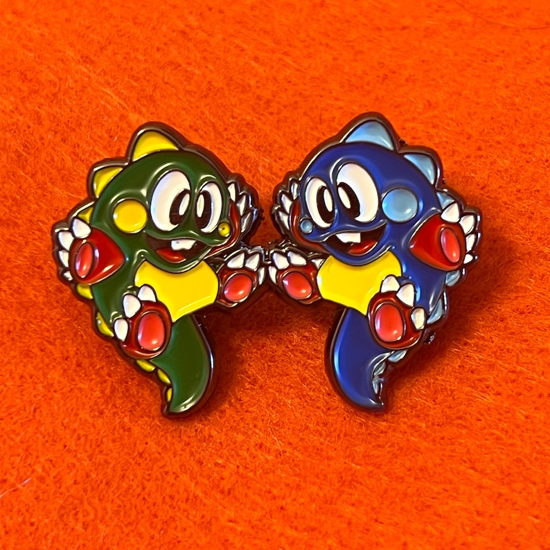 May include: Two enamel pins featuring cartoon dinosaur characters. One is green and yellow, the other is blue and yellow. Both have white eyes, red accents, and are smiling. The pins are set against an orange background.