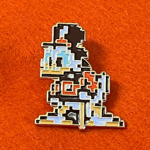 May include: A pixelated enamel pin featuring a cartoon character in a retro video game style. The pin has a black outline with blue, white, red, and tan details. The pin is set against an orange background.