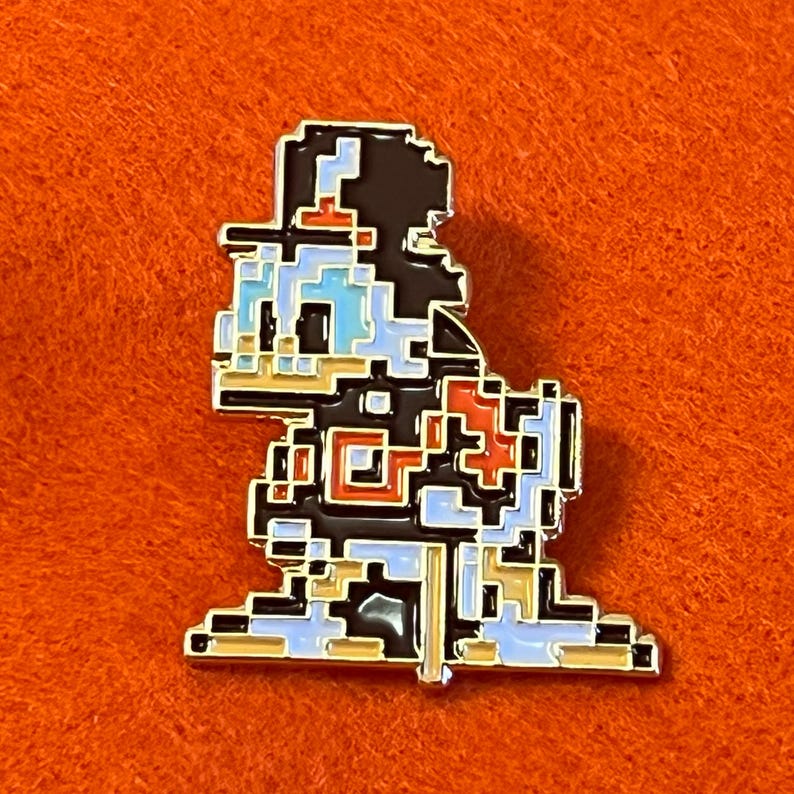 May include: A pixelated enamel pin featuring a cartoon character in a retro video game style. The pin has a black outline with blue, white, red, and tan details. The pin is set against an orange background.