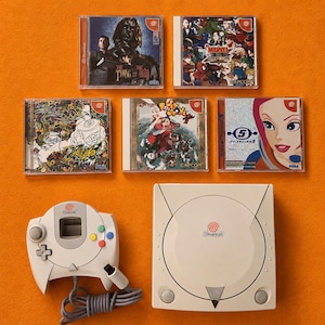 May include: A white Dreamcast console and controller are displayed with five game cases. The game cases feature various game titles and artwork. The console and controller are on an orange surface.