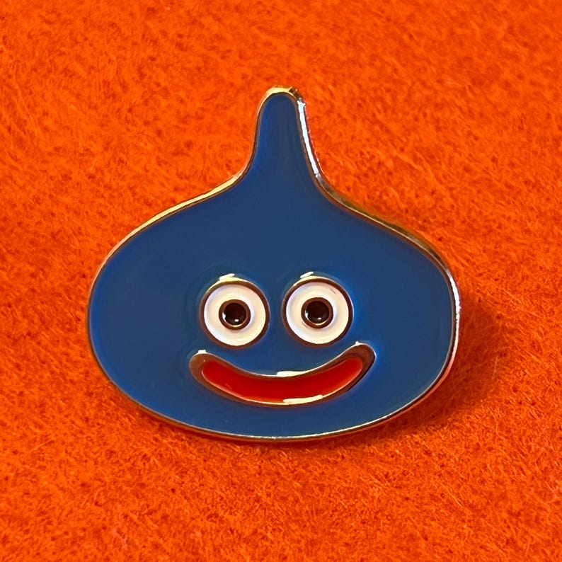 May include: A blue enamel pin in the shape of a smiling slime character. The pin has a gold-colored border, white eyes with black pupils, and a red smiling mouth. The pin is set against an orange background.