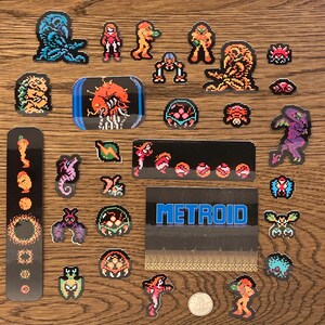 Może przedstawiać: Kolekcja naklejek w stylu pixel art z postaciami i elementami z gry wideo Metroid. Naklejki przedstawiają postacie w odcieniach pomarańczu, niebieskiego i fioletu, wraz ze słowem "METROID" w kolorze niebieskim.