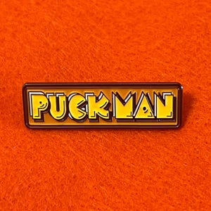 May include: A rectangular enamel pin with the word "PUCKMAN" in yellow, outlined in black and white, set against a brown background. The pin is on an orange background.