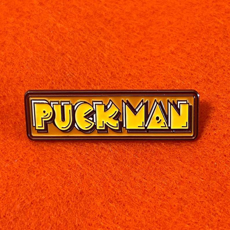 May include: A rectangular enamel pin with the word "PUCKMAN" in yellow, outlined in black and white, set against a brown background. The pin is on an orange background.