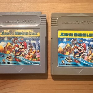 以下が含まれることがあります： 2つのグレーの任天堂ゲームボーイカートリッジ。どちらのカートリッジにも、マリオのカラフルなアートワークが施された「スーパーマリオランド」というゲームが収録されています。「1989 Nintendo」と「Made in Japan」の文字がカートリッジに表示されています。