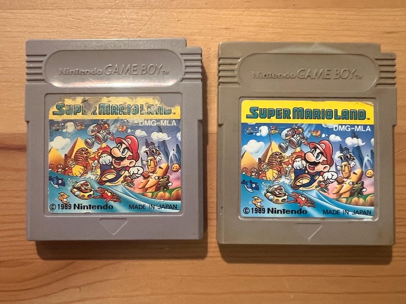 以下が含まれることがあります： 2つのグレーの任天堂ゲームボーイカートリッジ。どちらのカートリッジにも、マリオのカラフルなアートワークが施された「スーパーマリオランド」というゲームが収録されています。「1989 Nintendo」と「Made in Japan」の文字がカートリッジに表示されています。