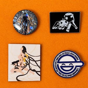 Op de afbeelding: Vier pins met anime-achtige illustraties op een oranje achtergrond. Eén ronde pin toont een figuur in een zwarte outfit. Een andere pin is rechthoekig met een wit robot-spin ontwerp. Een derde pin toont een vrouw met zwart haar. De laatste pin heeft een blauw-wit ontwerp met tekst.
