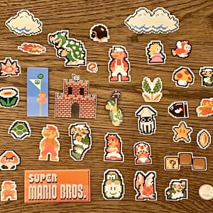 Może przedstawiać: Kolekcja pikselowych naklejek Super Mario Bros. z postaciami takimi jak Mario, Peach i Bowser, a także kultowymi elementami, takimi jak chmury, grzyby i bloki ze znakiem zapytania. Naklejki ułożone są na drewnianej powierzchni, z prostokątną naklejką z napisem "Super Mario Bros."