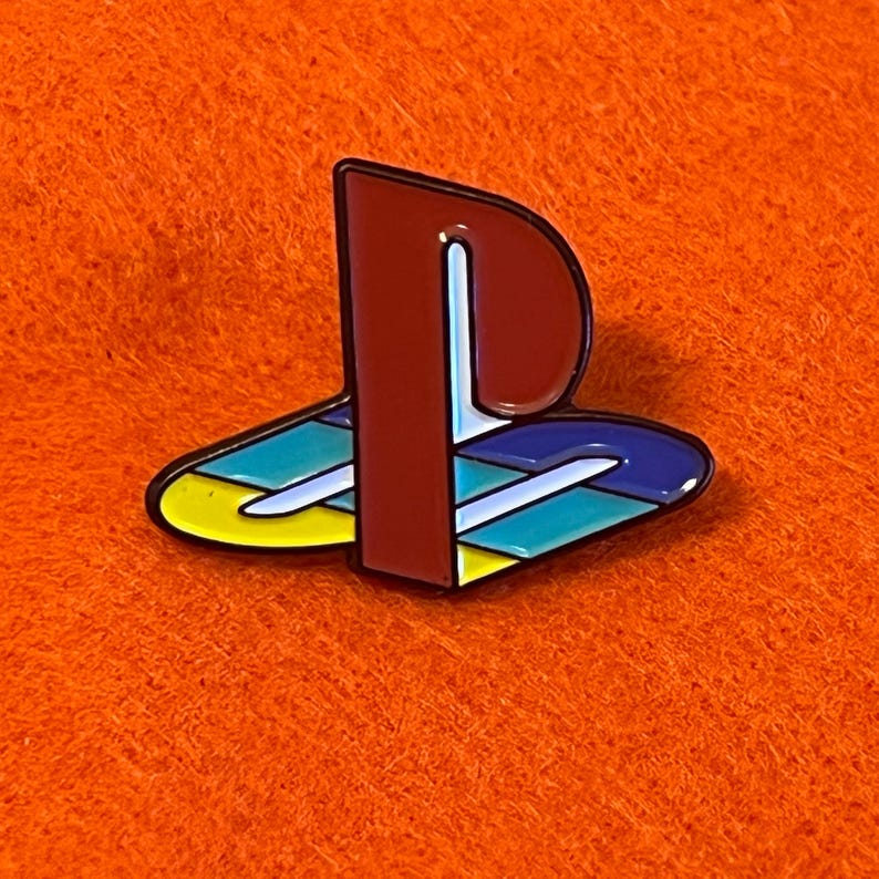 May include: A colorful enamel pin featuring the iconic PlayStation logo. The pin has a red and white 'P' shape over a stylized design with yellow, teal, and blue segments. The pin is set against an orange background.