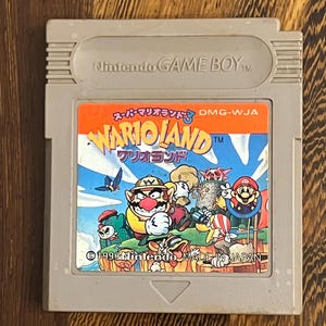 以下が含まれることがあります： Wario Landのグレーの任天堂ゲームボーイカートリッジ。ラベルには、ワリオや他のキャラクターのカラフルなイラストが描かれています。「WARIOLAND」と「Nintendo MADE IN JAPAN」の文字が含まれています。