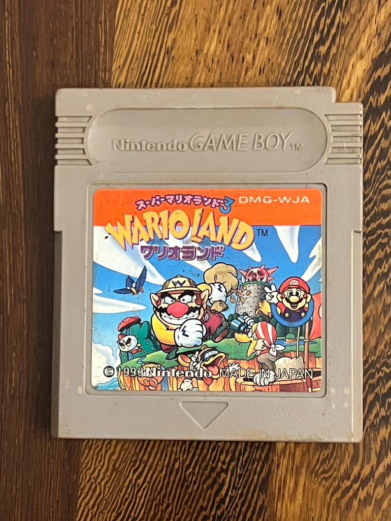 以下が含まれることがあります： Wario Landのグレーの任天堂ゲームボーイカートリッジ。ラベルには、ワリオや他のキャラクターのカラフルなイラストが描かれています。「WARIOLAND」と「Nintendo MADE IN JAPAN」の文字が含まれています。