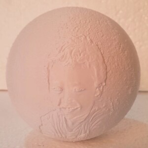 May include: White ceramic sphere with a 3D embossed portrait of a child. The child has short hair and is smiling.