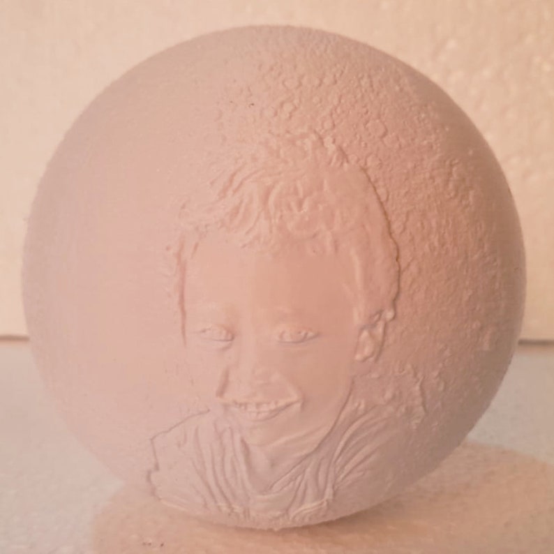 May include: White ceramic sphere with a 3D embossed portrait of a child. The child has short hair and is smiling.