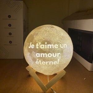 May include: A white moon-shaped night light with the words "Je t'aime un amour &eacute;ternel" inscribed on it. The light is on a wooden stand.