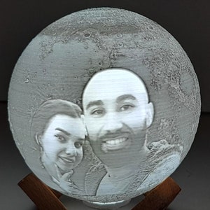 May include: A white 3D printed moon lamp with a couple's photo engraved on the surface. The lamp is on a wooden base.