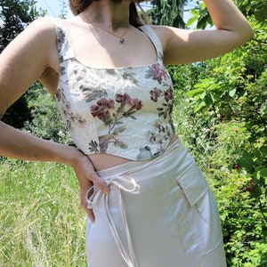 Peut inclure: Un haut court crème avec un motif floral dans les tons bordeaux et vert. Le haut est associé à une jupe portefeuille crème assortie. La tenue est mise en valeur sur un fond de verdure et de soleil.