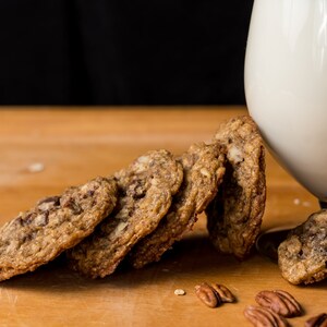 May include: A close-up of a stack of oatmeal chocolate chip cookies on a wooden surface. The cookies are golden brown and have a slightly crispy texture. There is a glass of milk in the background.