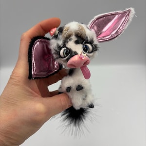 Peut inclure: Une vache en peluche faite à la main avec un motif tacheté noir et blanc, de longs cils et des accents roses sur les oreilles et la langue. La vache a une queue touffue.