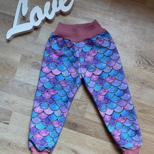 Op de afbeelding: Broek met een zeemeerminstaartpatroon in blauw, paars en roze. De broek heeft een hoge tailleband en manchetten in effen roze. Het woord "Love" staat in witte letters op de achtergrond.