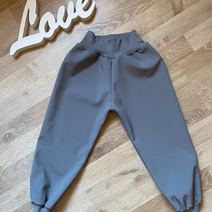 Op de afbeelding: Grijze peuterbroek met een hoge tailleband en manchetten. De broek is gemaakt van een zachte, gladde stof. Een wit houten bord met de tekst "Love" staat op de achtergrond.