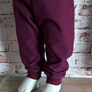 Könnte beinhalten: Pflaumenfarbene Babyhose mit breitem, weinrotem Bund und passenden Bündchen. Die Hose ist aus einem weichen Stoff gefertigt und hat eine lockere Passform. Die Hose wird auf einem weißen Schaufensterpuppenfuß präsentiert.