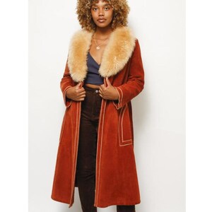 May include: A long, rust-coloured coat with a faux fur collar. The coat features cream-coloured stitching along the edges and a tailored fit. The model is wearing a navy blue top and brown trousers. The coat is a stylish outerwear option.